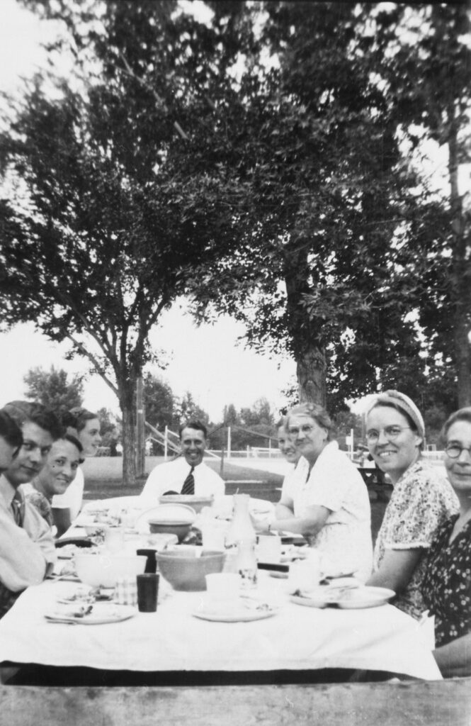 Women sitting around a picnic table with a man at the head with trees in the background