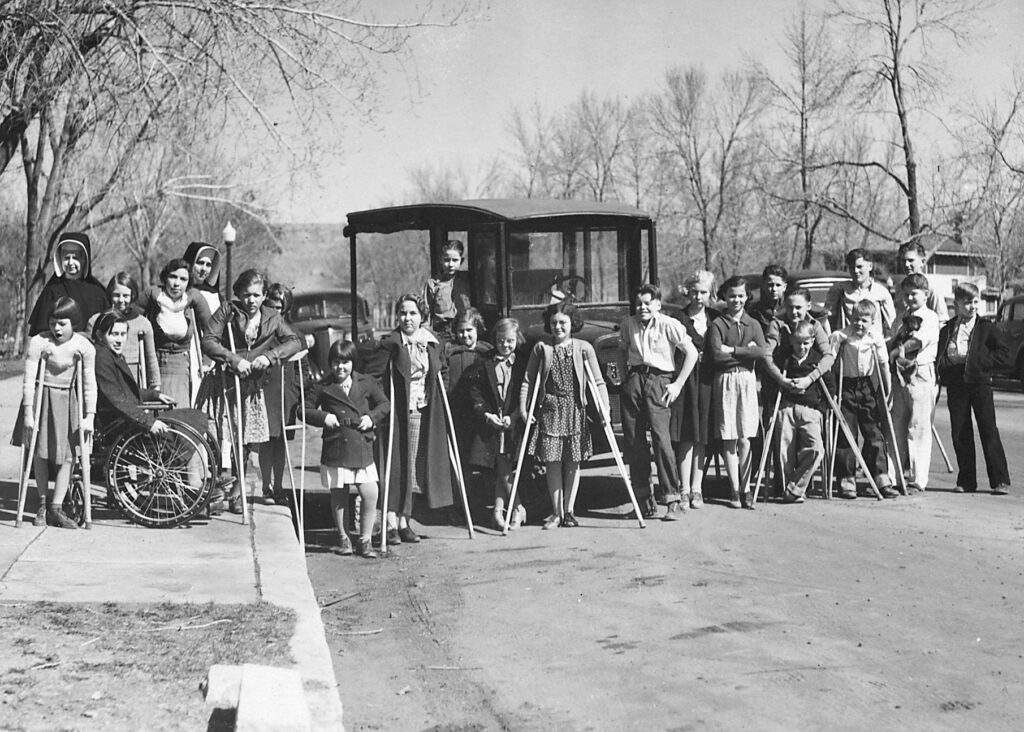 Group of children standing in front of a old car. Children are using a variety of crutches, leg braces, and wheelchairs