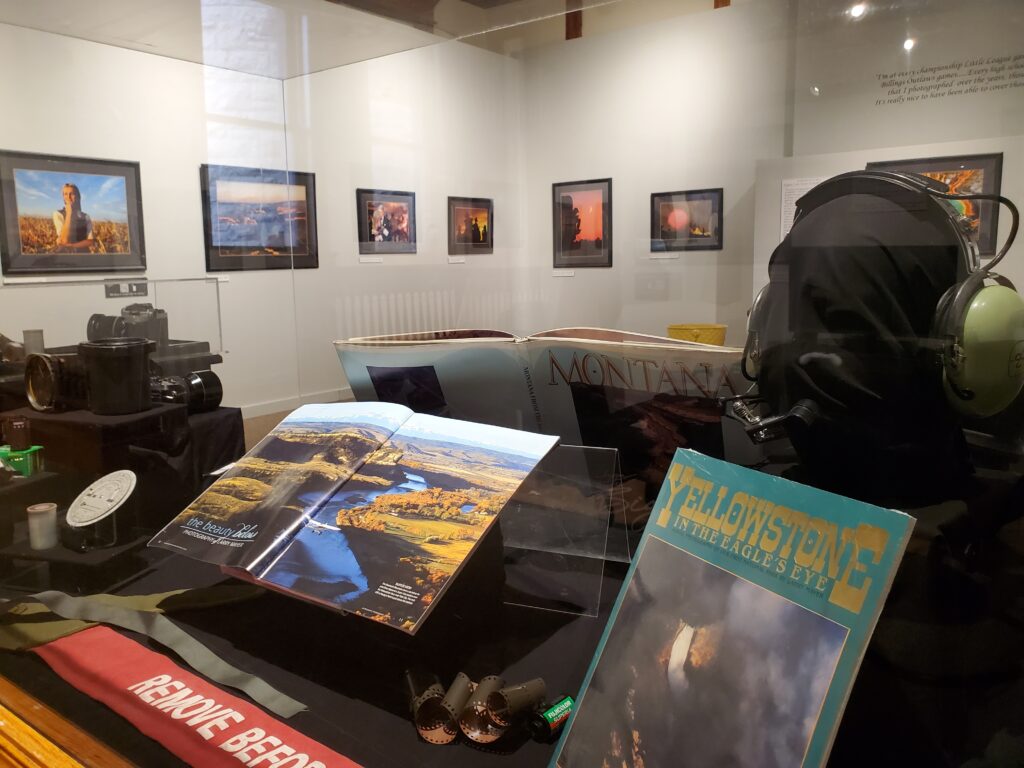 Books, photography lenses, and headphones displayed behind glass with framed photographs in the background