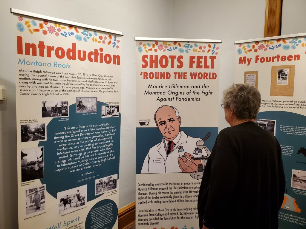 An older woman looks at pop up banners with an illustrated image of a scientist and microscope. Banner says "Shots Felt Round Montana"